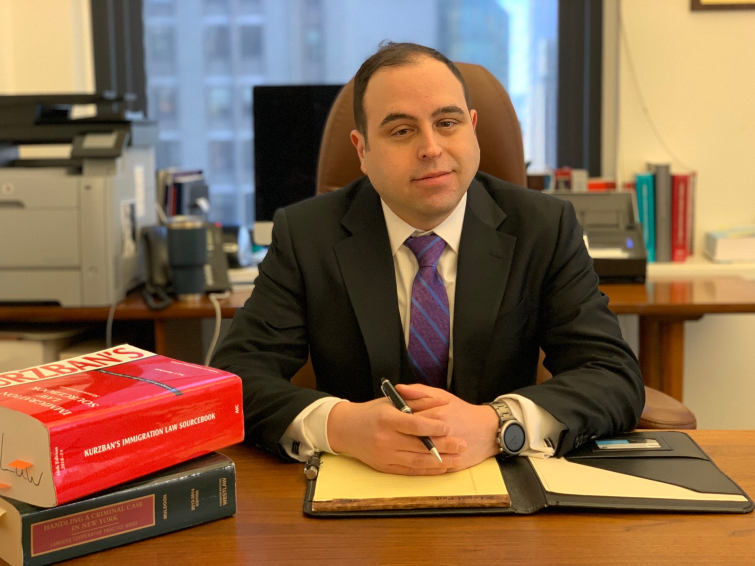Attorney Lee A. Koch sitting at a desk with his hands folder wearing a black suit and purple and blue striped tie. With a red bound "Kurzban's Immigration Sourcebook" and a blue bound "Handling a Criminal Case in New York" to his right and a yellow legal notepad under his hands.